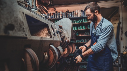 Bärtige Person mit Brillen in Arbeitsschürze steht an großer Poliermaschine und hält Lederschuh an Riemen, im Hintergrund Regale mit Spraydosen und Schuhmodeln
