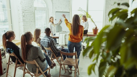 Personen sitzen und stehen in einem Raum mit Flipchart und tauschen sich aus