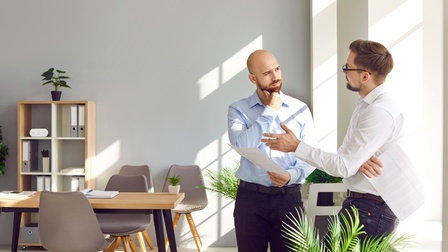 Zwei Männer in Hemden verhandeln stehend im Büro an einer Fensterbank