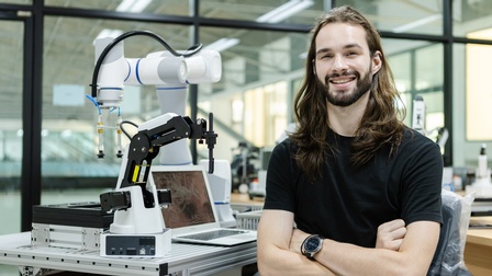 Lächelnde Person mit Bart und langem Haar im Portrait hält Arme vor Körper verschränkt, im Hintergrund Tisch mit aufgeklapptem Notebook und Roboterarm
