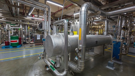 In einer Produktionsstätte stehen verschiedene große Maschinen mit Rohren und Verbindungen.