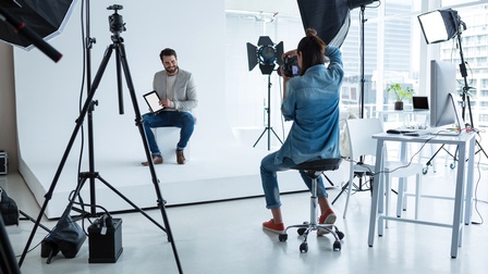 In einem Fotostudio sitzt eine Person vor einem weißen Hintergrund auf einem weißen Hocker. Die Person hält ein Tablet und lächelt. Ihr gegenüber sitzt eine Person, die mit einer Kamera Fotos macht, ringsum stehen Stative mit Lightboxen und Scheinwerfen.