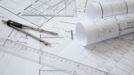 Nahaufnahme mehrerer, übereinander liegender Papiere, auf denen Pläne mit Grundrissen sind. Auf den Papieren liegen zwei Papierrollen, zwei Lineale und ein Zirkel.