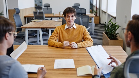 An einem Tisch sitzen an einer Seite zwei Personen. Die rechte Person hält ein Klemmbrett. Vor beiden Personen liegen aufgeklappte Notizbücher. Ihnen gegenüber sitzt eine weitere Person