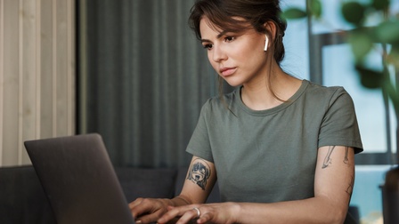 Person mit Earpods, kurzem grünen T-Shirt und Tattoos sitzt an einem Schreibtisch und tippt konzentriert in einen Laptop
