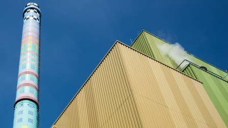 Außenansicht eine Biomasse-Kraftwerks unter blauem Himmel: Bunter Turm neben Gebäude mit gelber und grüner Blechverkleidung