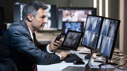 Nahaufnahme einer Person, die in einem Bildschirm eines Computers sitzt, in dem Videoaufnahmen eines Ganges zu sehen sind. Sie tippt auf einer Tastatur, die auf einem Schreibtisch liegt, in der linken Hand hält sie ein Walkie Talkie. 