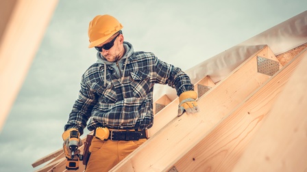 Person mit gelben Schutzhelm, Sonnenbrille und karierter Jacke steht in einem Dachausbau aus Holz