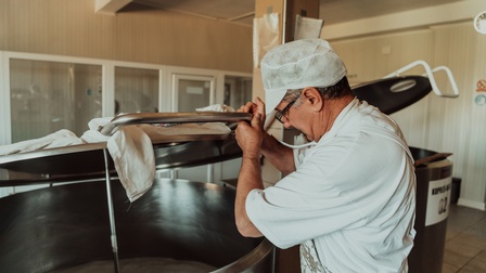 Eine Person mit Haarnetz steht neben einem großen, metallenen Behälter, in dem Joghurt drinnen ist. Die Person hebt den Deckel des Behälters an