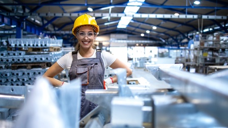 Lächelnde Person in Arbeitskleidung mit gelben Schutzhelm und Schutzbrille steht in einer industriellen Produktionsstätte