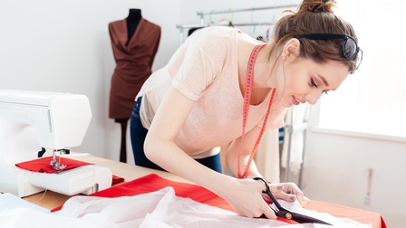 Person mit Maßband um den Hals schneidet Stoff über Tisch gebeugt, daneben Nähmaschine, im Hintergrund Kleiderpuppe