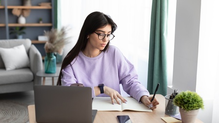 Person mit langen dunklen Haaren und Brille sitzt bei einem Schreibtisch mit Laptop und notiert sich etwas in einem Notizbuch