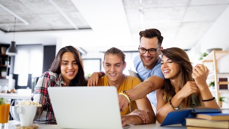 Gruppe von jungen Erwachsenen blickt freudig auf einem Laptopbildschirm in einer modernen Büroräumlichkeit