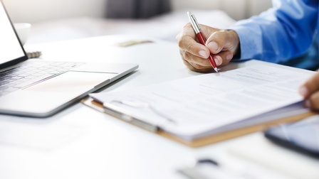 Eine Person sitzt an einem Schreibtisch. Vor ihr liegt ein Klemmbrett, auf dem Zettel sind. Die Person hält einen Stift. Neben dem Klemmbrett ist ein Laptop