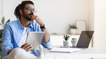 Eine Person sitzt an einem Tisch und blickt nachdenklich in die Ferne. Am Tisch stehen ein Laptop, eine Kaffeetasse und ein Notizbuch. Die Person hält ein Tablet in der Hand.