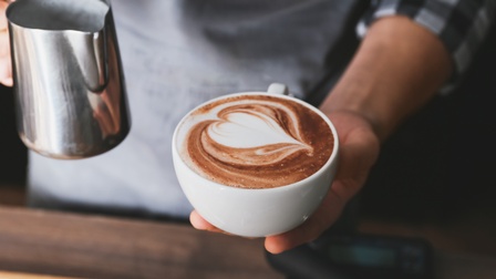Detailansicht einer Person, die Kaffeetasse gefüllt mit Kaffee und Milchschaum in einer Hand hält, in anderer Hand Edelstahlmilchkanne