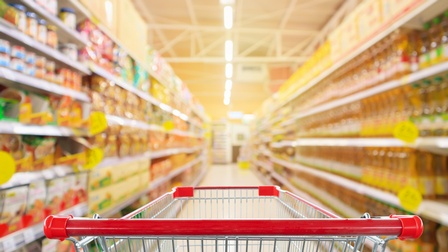 Aufnahme eines leeren Einkaufswagens, der im Gang eines Supermarkts steht. In den Regalen sind verschiedene Produkte und Lebensmittel