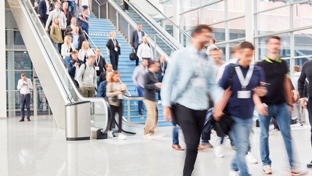 Im Vordergrund gehen Personen mit Ausweisen um den Hals. Im Hintergrund sind eine Treppe sowie eine Rolltreppe mit weiteren Personen, die hinunter und hinaufgehen