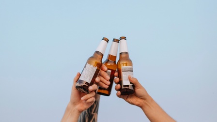 Drei nach oben gestreckte Hände vor blauem Hintergrund, die jeweils eine Bierflasche halten und miteinander anstoßen