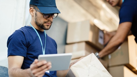 Eine Person mit Kappe, Brille, Bart und Ausweis um den Hals hält in der rechten Hand ein kleines Tablet. In der linken Hand hält sie einen Karton. Im Hintergrund sortiert eine andere Person Kartons