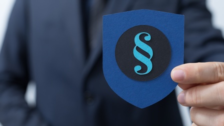 Nahaufnahme der linken Hand einer Person, ansonsten ist nur der Oberkörper ohne Gesicht verschwommen zu sehen. Sie hält ein blaues Papier in Wappenform in der Hand, auf dem ein schwarzer Kreis klebt. Auf diesem klebt ein türkises Paragraphen-Zeichen. 