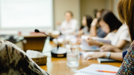 In einem Raum sitzen mehrere Personen um einen Tisch. Vor den Personen sind teilweise Unterlagen und Stifte. Auf dem Tisch steht ein Mikrofon. Im Hintergrund ist ein großer Bildschirm an der Wand