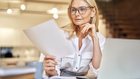 Eine Person mit Brille und in einer Bluse sitzt an einem Tisch in einem Wohnessbereich. Die Person hält einen Zettel, auf den sie blickt. Vor ihr steht ein aufgeklappter Laptop. Im Hintergrund sind eine Küche und eine Treppe