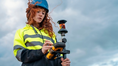 Person mit Helm in Schutzbekleidung steht unter blauem Himmel, Wind weht durch Haar und blickt auf Messgerät, das sie in Händen hält.