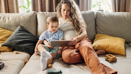 Auf einem tiefen Sofa sitzt ein Kind neben einer erwachsenen Person mit einem nach außen gewölbten Bauch, die ein Tablet in den Händen hält, auf das Kind und erwachsene Person blicken. Auf dem Sofa liegen Spielsachen