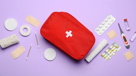 Eine rote Erste Hilfe Tasche mit weißem Logo liegt gemeinsam mit Medikamenten, Verbandsmaterial, Ampullen, Wattestäbchen, Wattepads sowie einem Fiebermesser auf einem lila Untergrund, Topshot