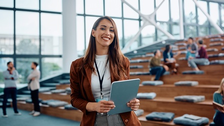 Halbporträt einer Person mit einem Tablet in beiden Händen sowie einem Ausweisschild um den Hals. Hinter der Person sind mehrere sich stufenweise erhöhende Holzbänke, auf denen teilweise graue Kissen liegen