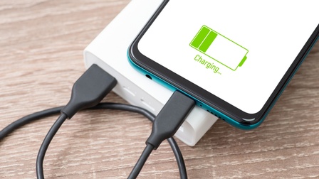 Nahaufnahme des unteren Teils eines Smartphones, auf dessen Bildschirm ein grüner Akku mit zwei Balken ist, unter dem das Wort Charging steht. Das Smartphone ist mit einem schwarzen Kabel verbunden, das mit einer weißen, kleinen Box verbunden ist.