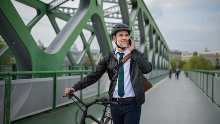 Person mit schwarzem Fahrradhelm schiebt Rad über Brücke mit grüner Stahlkonstruktion und telefoniert