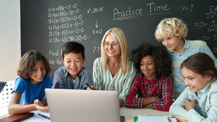 Lehrperson sitzt mit Schüler:innen vor aufgeklappten Laptop, im Hintergrund beschriebene Kreidetafel