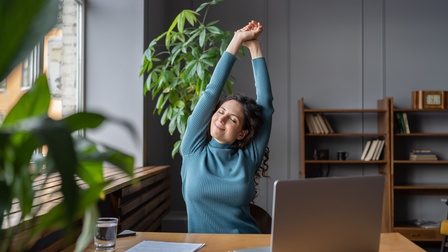 Eine Person in einem bläulichen Oberteil sitzt an einem Tisch. Die Person hat die Augen geschlossen und streckt ihre beiden Arme nach oben. Auf dem Tisch ist ein aufgeklappter Laptop. Im Hintergrund ist ein Regal mit Büchern