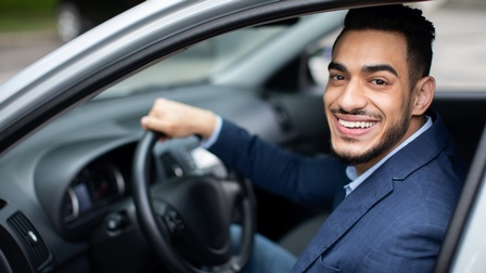 Lächelnde Person in Businesskleidung mit dunklen kurzen Haaren und Bart sitzt freudig am Steuer eines Autos