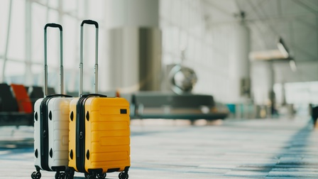 Ein grauer und ein gelber Handgepäckskoffer stehen auf Teppichboden in einem langen Gang mit großen Fensterfronten und Sitzmöglichkeiten