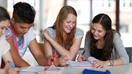 Lächelnde junge Personen an Tisch sitzend auf Heft mit Notizen blickend, in Händen Stifte haötend