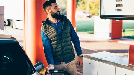 Eine Person steht am rechten hinteren Ende eines Autos und hält mit der rechten Hand eine Zapfpistole in eine Öffnung des Autos. Die Person blickt auf eine Zahlenanzeige, die in Höhe ihres Kopfes hängt.