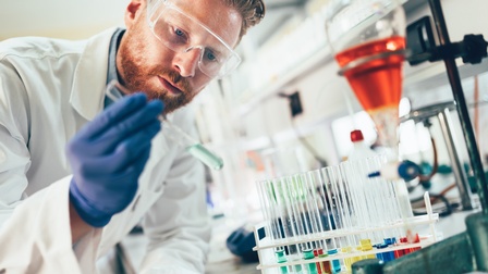 Person mit kurzen Haaren und Bart trägt eine weiße Schutzkleidung mit Brille und blauen Handschuhen und blickt auf ein Reagenzglas mit Flüssigkeit in der Hand während im Hintergrund weitere Reagenzgläser mit Flüssigkeiten in einem Labor stehen