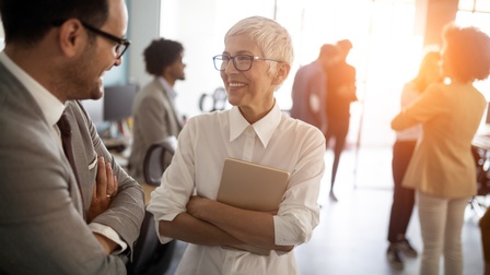 Lächelnde Person mit Brillen hält Tablet in verschränkten Armen und blickt auf andere mit ihr im Gespräch befindliche Person in Seitenansicht, im Hintergrund verschwommen weitere Personen