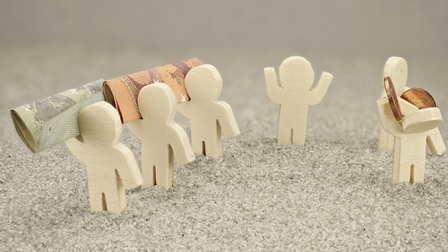 Nahaufnahme eines hellen Bodens, auf dem sechs kleine Figuren von Personen aus einem hellen Material sind. Drei Figuren tragen Geldscheine. Zwei Figuren tragen Münzen. Eine dieser Figuren hat einen nach unten geneigten Kopf