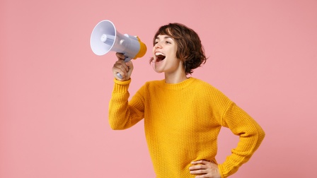 Person mit kurzen braunen Haaren, Ohrringen und gelben Pullover hält ein Megaphon und verlautbart etwas vor einem rosa Hintergrund