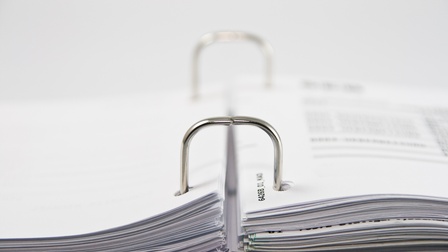 Nahaufnahme eines aufgeklappten Spiralordners mit Papierdokumenten vor einem weißen Hintergrund