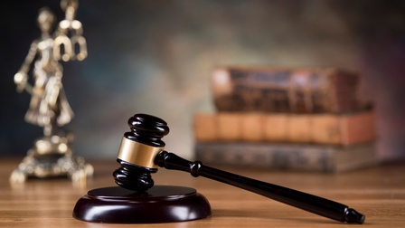 Ein Richterhammer liegt auf einem Holzuntergrund, im Hintergrund steht eine Justitia Statue sowie Bücher in der Unschärfe