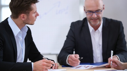 Zwei Personen in Jackets an Tisch sitzend, auf dem Unterlagen mit Diagrammen liegen