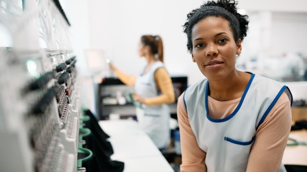 Sanft lächelnde Person in hellblauer Arbeitskleidung steht vor industrieller Stickmaschine, im Hintergrund verschwommen weitere Person