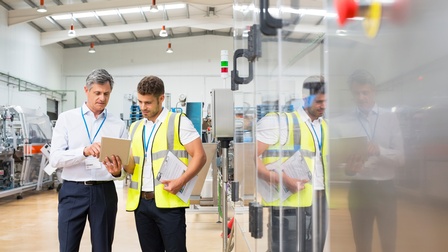 Person mit Businesskleidung und Ausweiskarte zeigt auf ein Tablet und eine zweite Person mit Warnweste und Klemmbrett blickt aufmerksam drauf in einer technischen Betriebsstätte