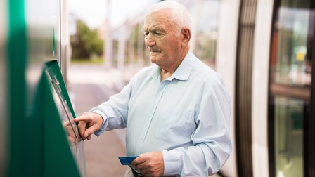 Ältere Person mit weißen kurzen Haaren und hellblauem Hemd steht bei einem Bankomat und hält eine Bankomatkarte bereit