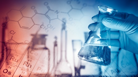 Fokus auf Laborglas, das Hand in Gummihandschuh hält, im Hintergrund Schatten von weiteren Laborgefäßen und Ausschnitte des Periodensystems und molekulare Ketten